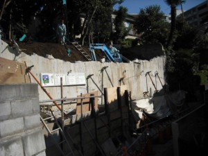 下部の施工はＨ鋼を打込み矢板にて山留。上部に行くにつれ、間知石ブロックの裏込めコンクリートが擁壁にあたってきました。 　　上部よりカニクレーンで土砂を上げ、ハンドクラッシャーにて擁壁を壊しながら少しずつ地盤を下げ、中段と下段にバックホウを配置しての作業です。